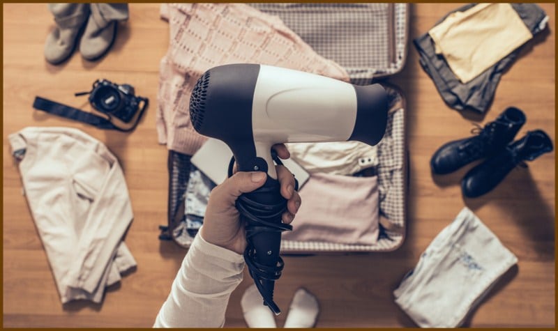 A woman holds a hairdryer & packing clothes in a suitcase for traveling