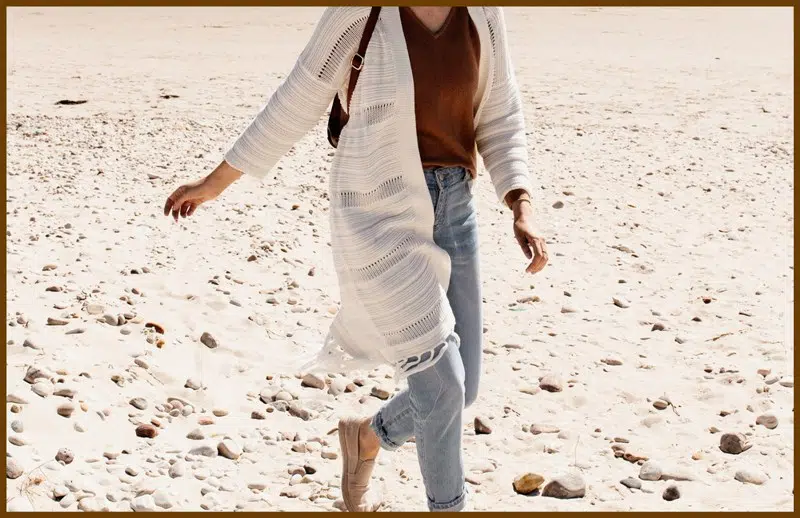 A woman is walking on a sandy beach with a cardigan